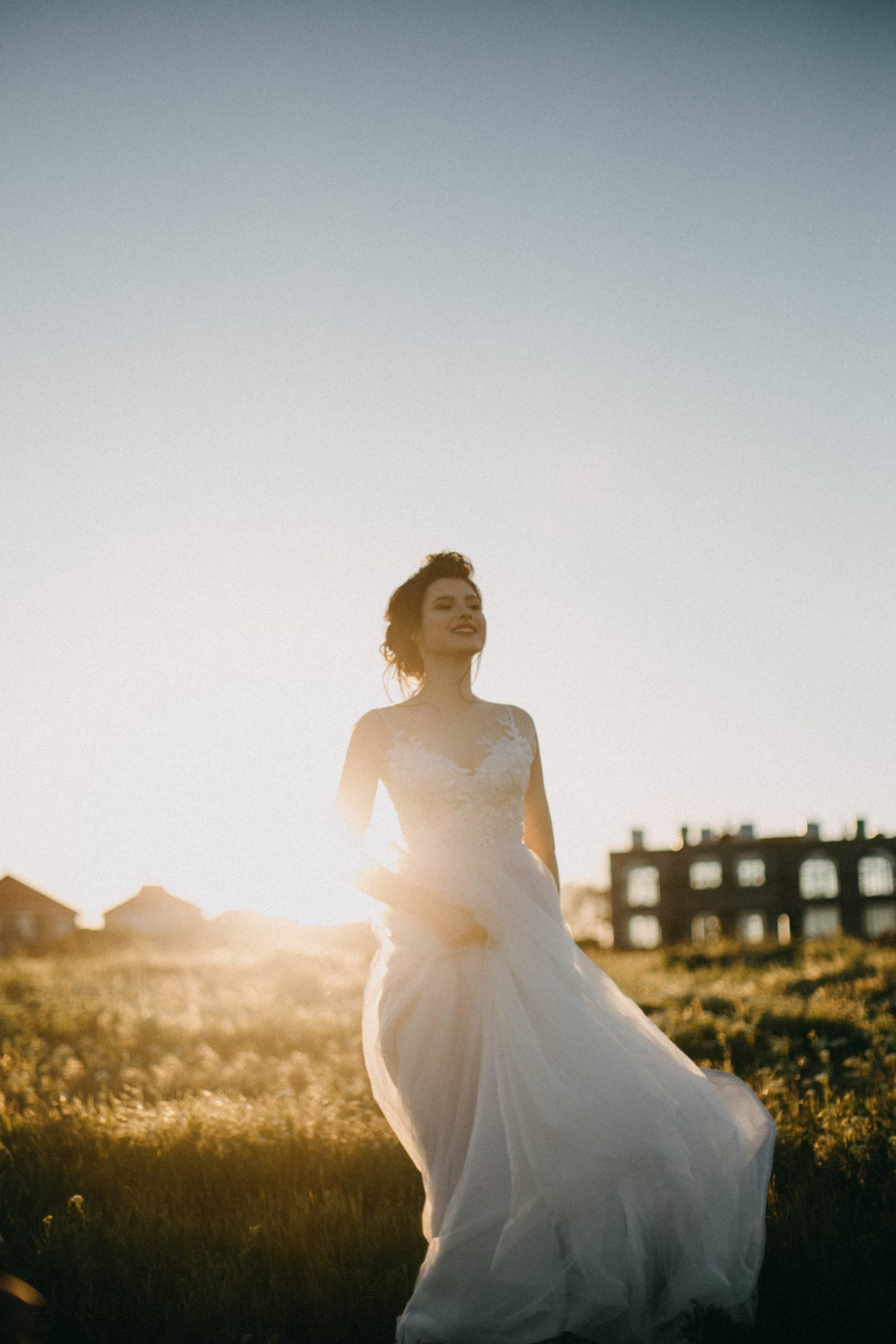A beautiful bride with a white gown and veil stands outside on a sunny and windy day, smiling happily.