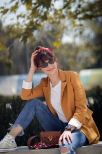 Image of a happy woman sitting and wearing sunglasses with a jacket that has been customized by Rose N Silk. The jacket has intricate designs and textures, and complements the woman's outfit. The woman appears relaxed and content, enjoying her surroundings.